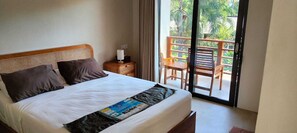 A hotel room with a bed, a nightstand, a window with a view of greenery, and a balcony with wooden furniture. - Trawangan Oasis (Gili Trawangan)