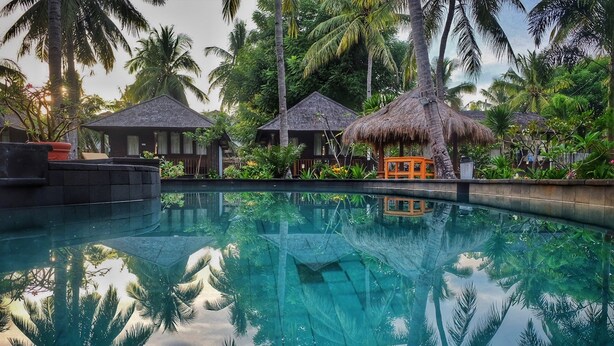 A pool with reflections of palm trees and thatched-roof structures, surrounded by lush greenery. - Trawangan Oasis (Gili Trawangan)