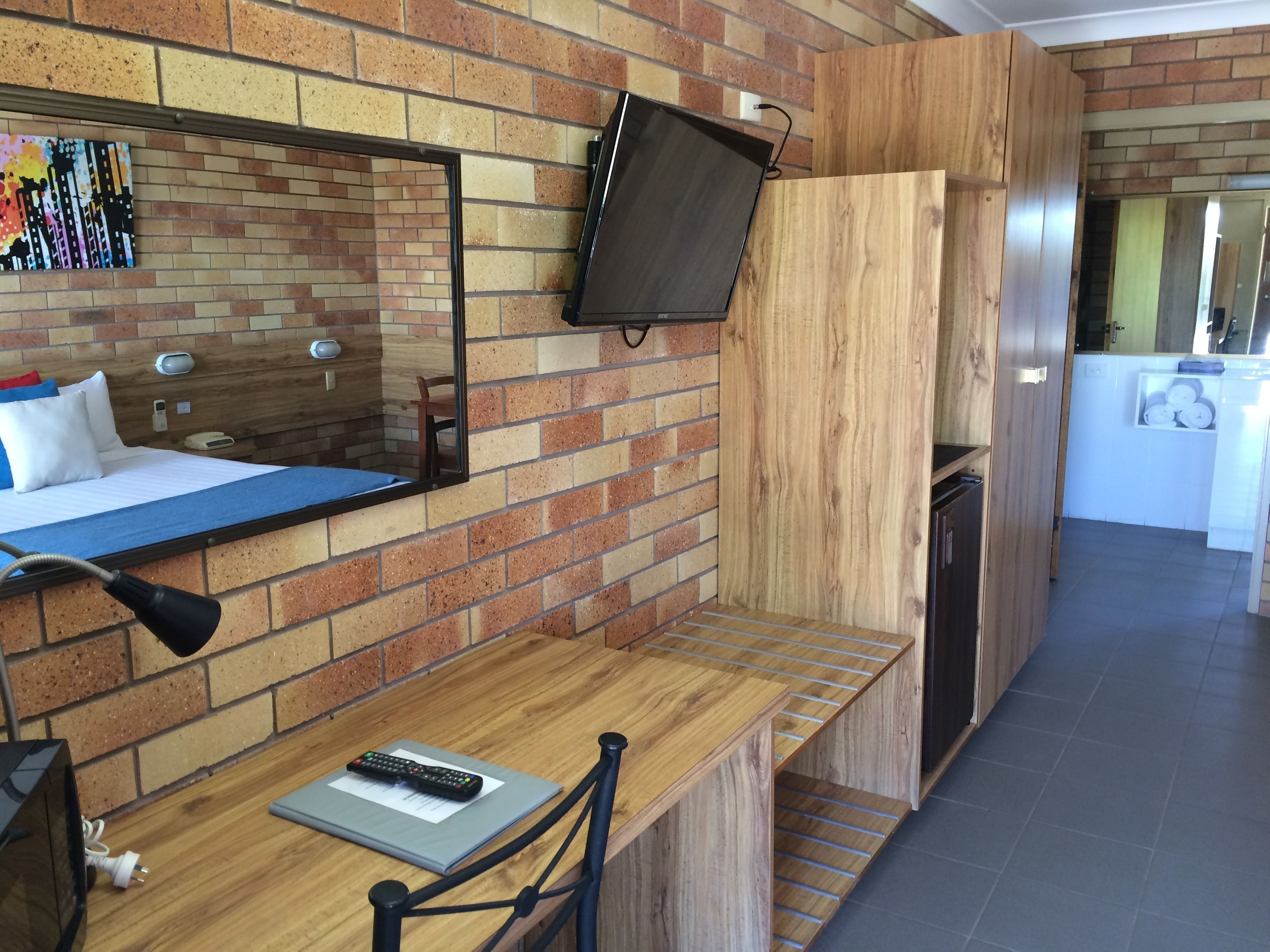 Quarto de hotel com parede em padrão de tijolos, cama com roupa de cama azul, TV montada, escrivaninha com controle remoto e um espelho.