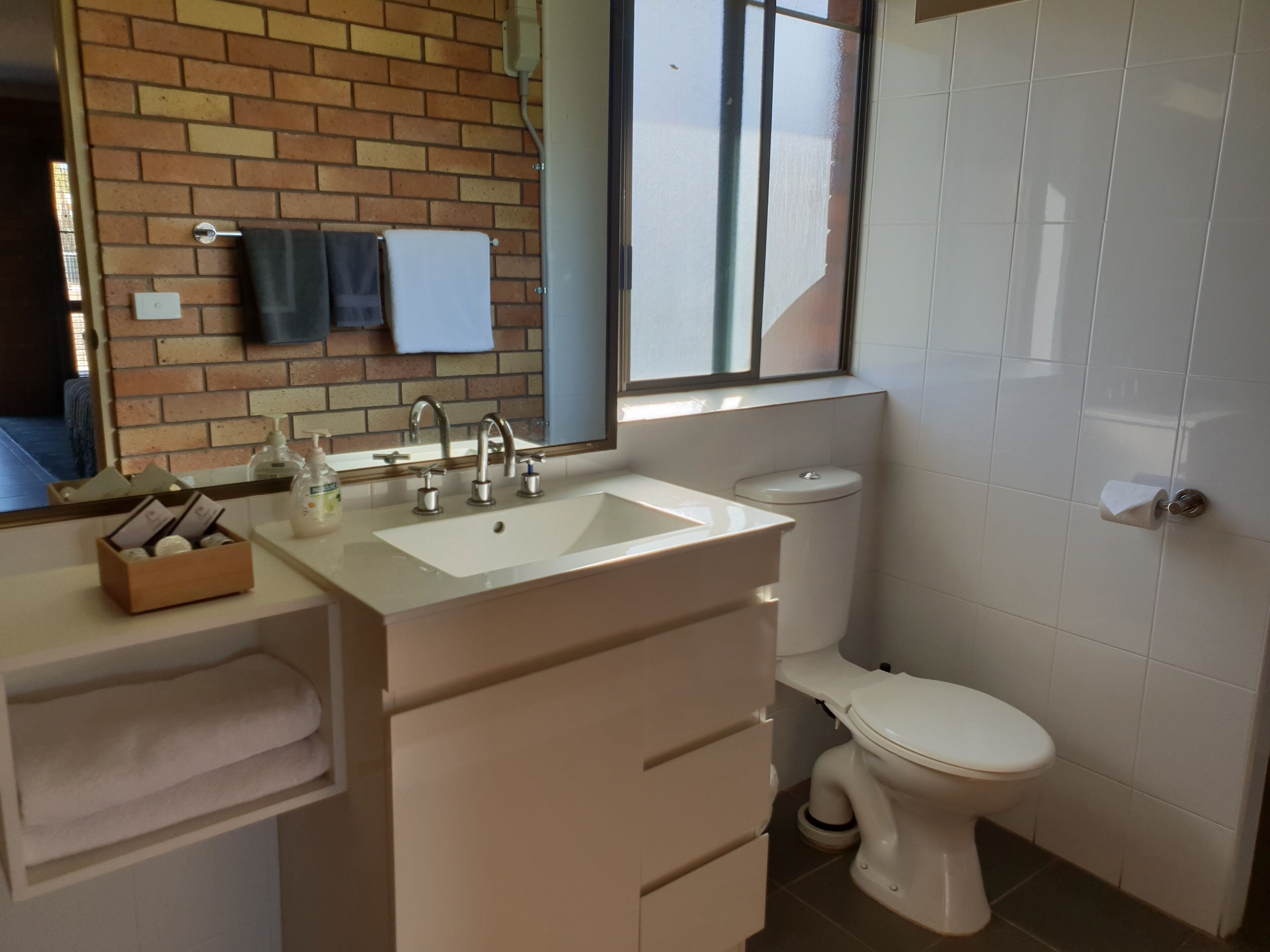 A bathroom with a white sink, a toilet, and a mirror. There is a window above the sink and a towel rack on the wall.