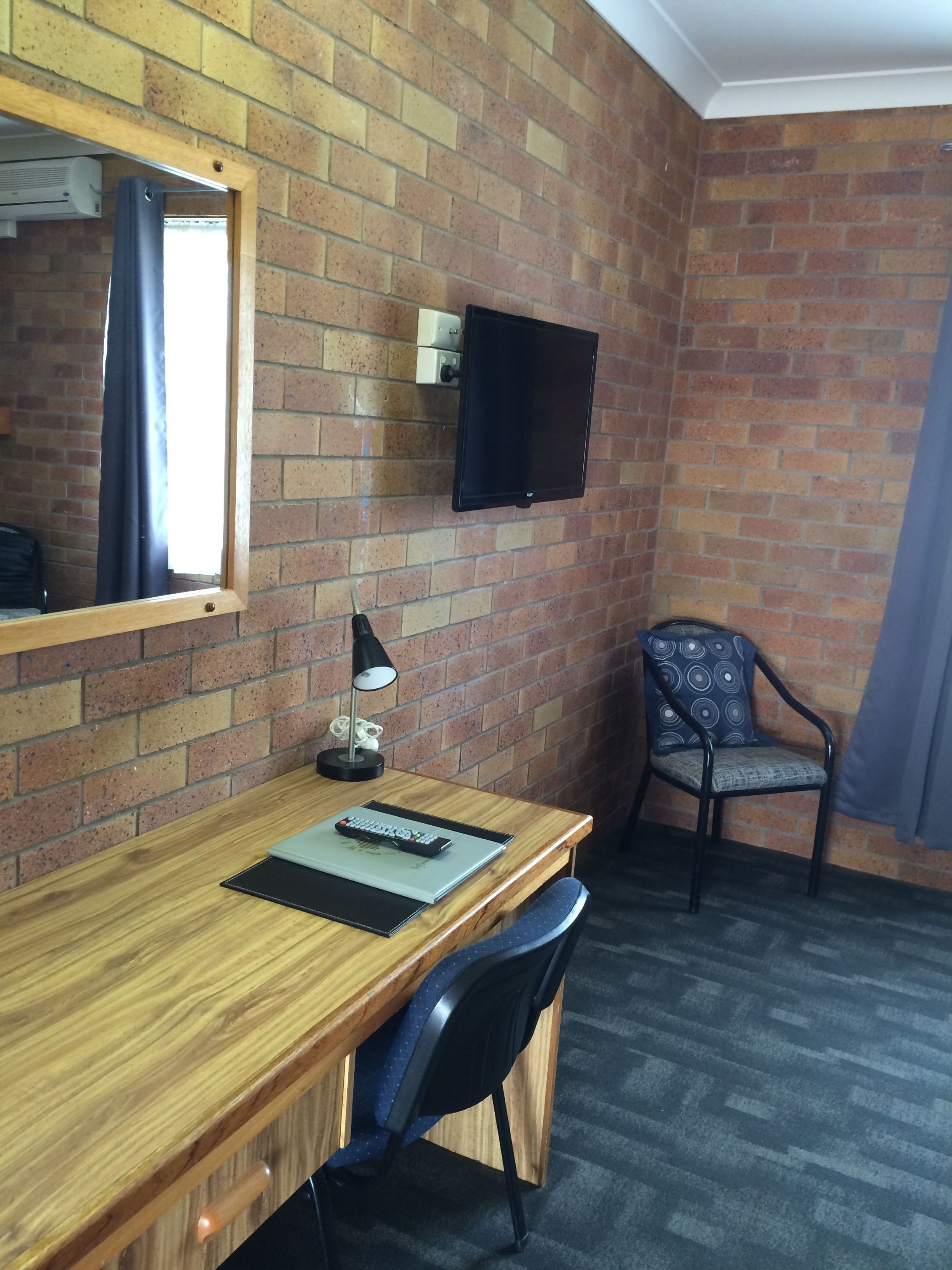 A hotel room with a wooden desk, a chair, a TV mounted on the brick wall, and a window with curtains.