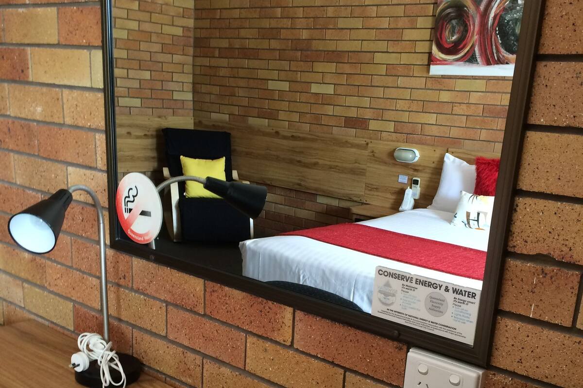 A hotel room with a brick wall, a bed with a red and white bedspread, a black chair, a yellow pillow, a white bedside table, a lamp, a remote control, and a power outlet.