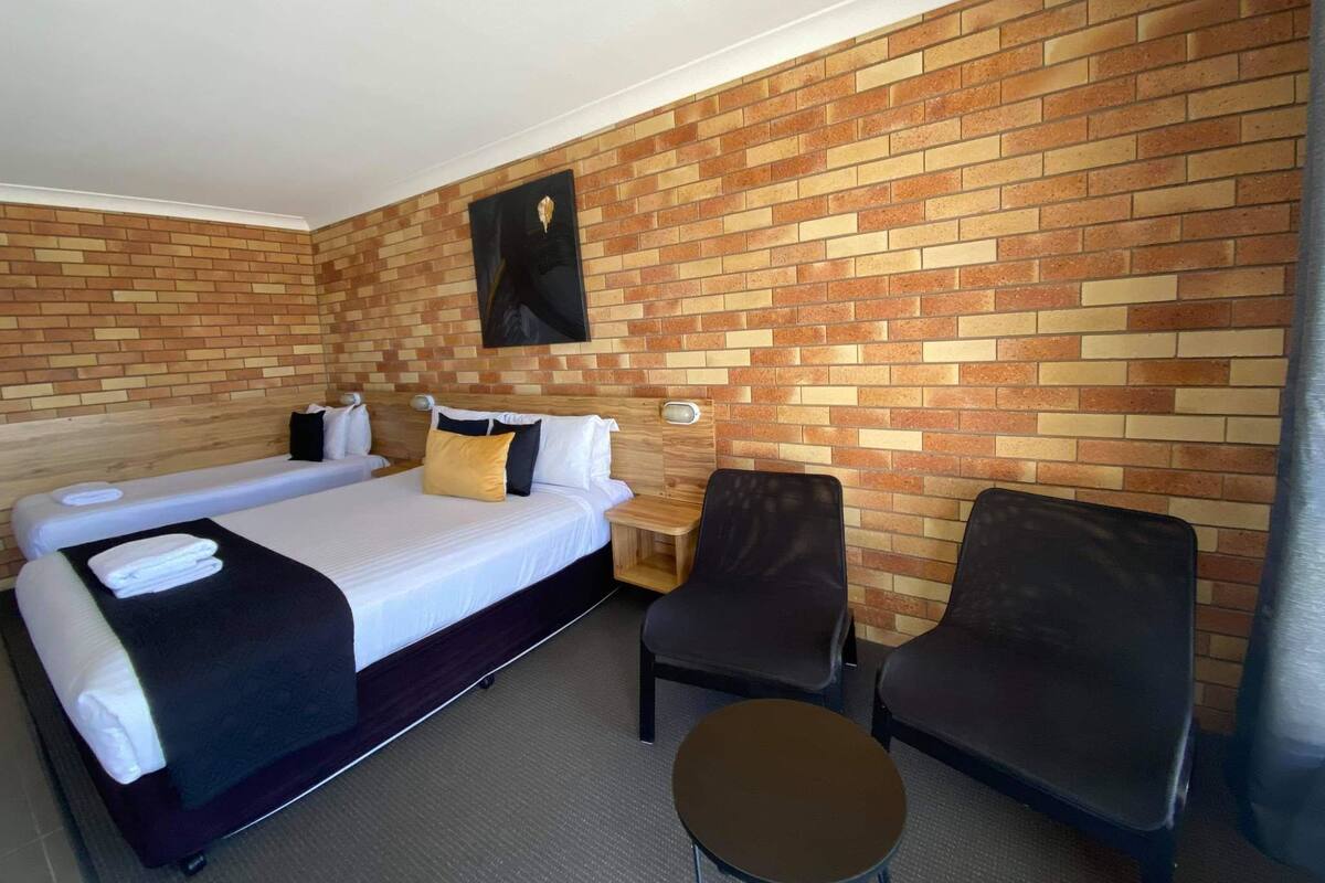 A hotel room with a brick-patterned wall, a bed with white linens, two black chairs, and a small round table.