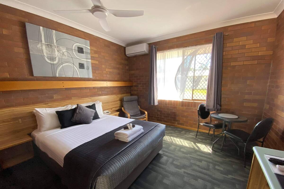 A hotel room with a large bed, a desk, a chair, and a small table. There is a ceiling fan, a window with curtains, and a brick wall.