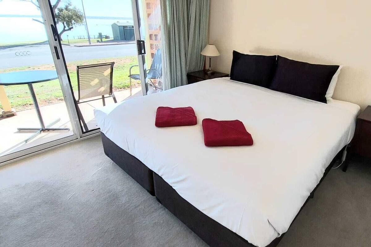 A hotel room with a large bed, two red pillows, a nightstand with a lamp, and a view of the outdoors through a glass door.