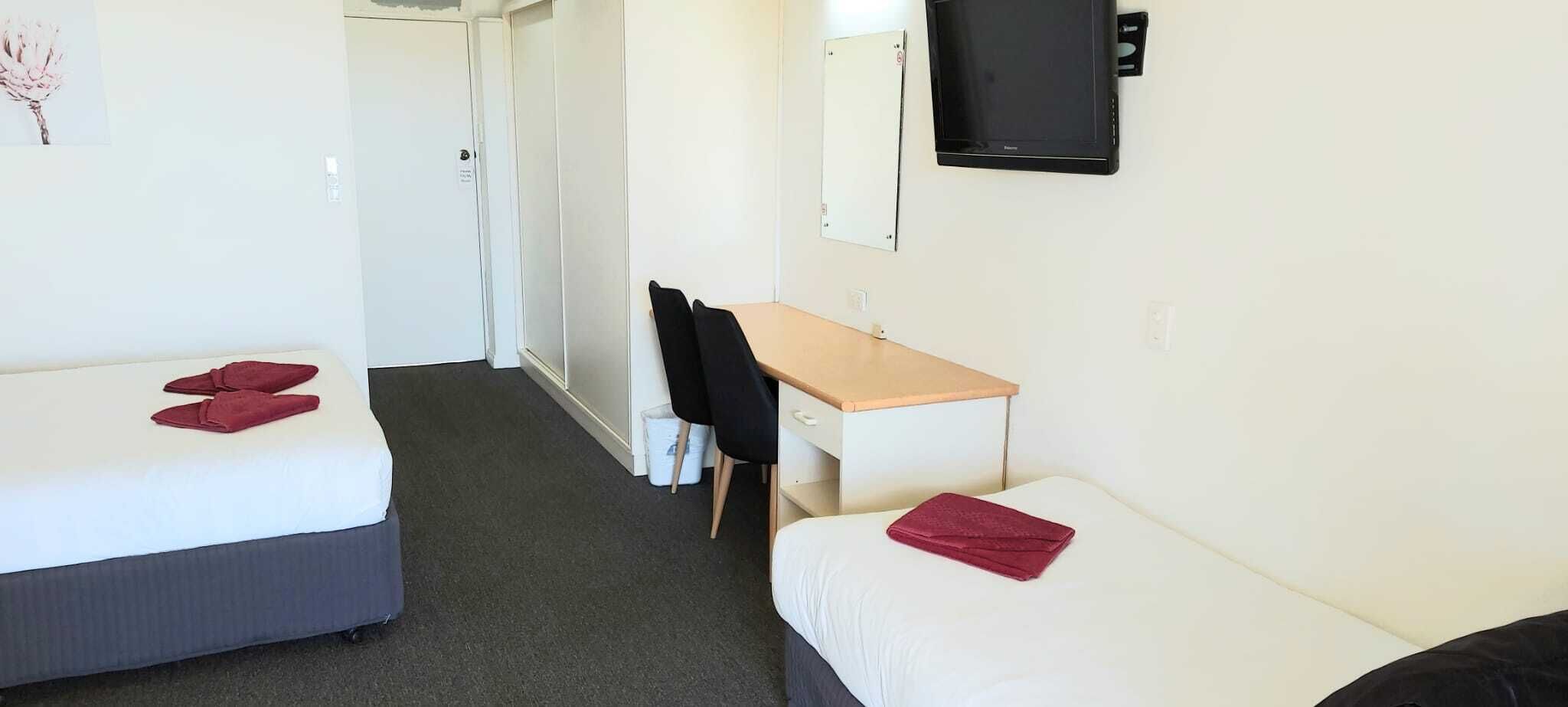 A hotel room with two single beds, a desk, a chair, a TV, and a trash bin.