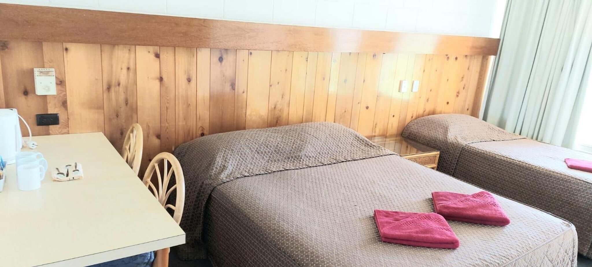 A room with two single beds, a wooden headboard, a table with a white mug, and a chair.
