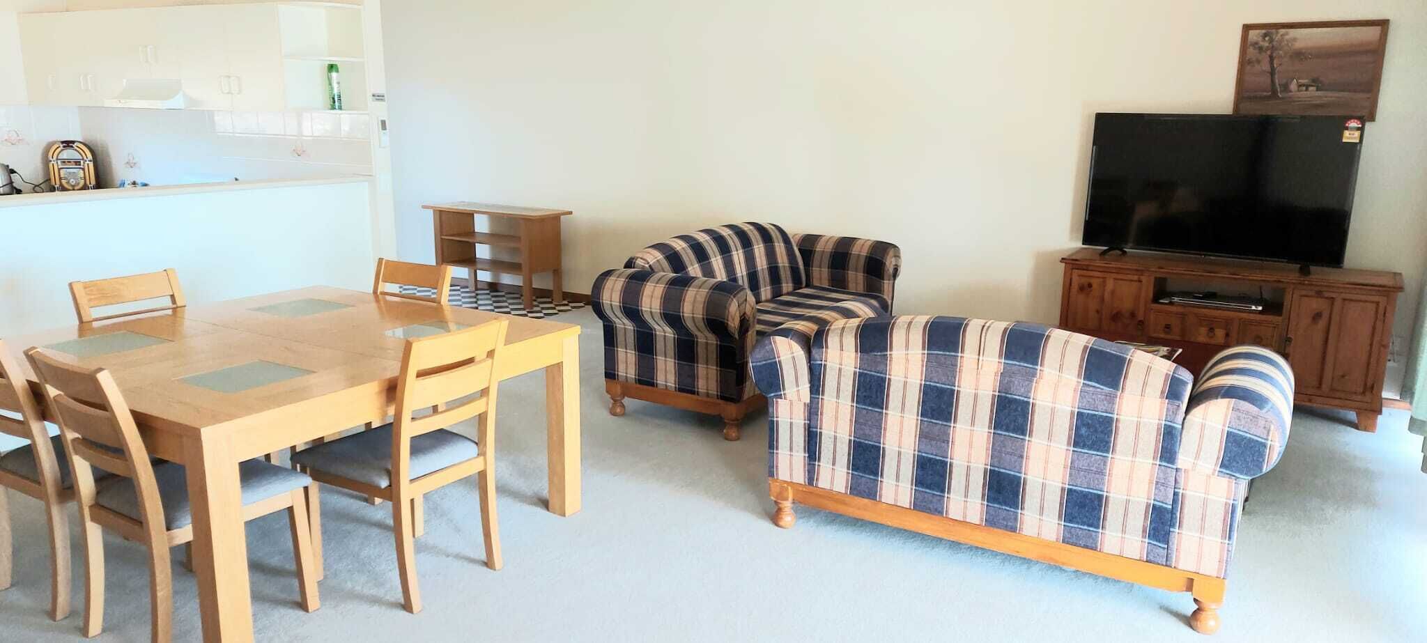 Una sala de estar con una mesa de comedor de madera, sillas, un sofá a cuadros, un televisor y un aparador.
