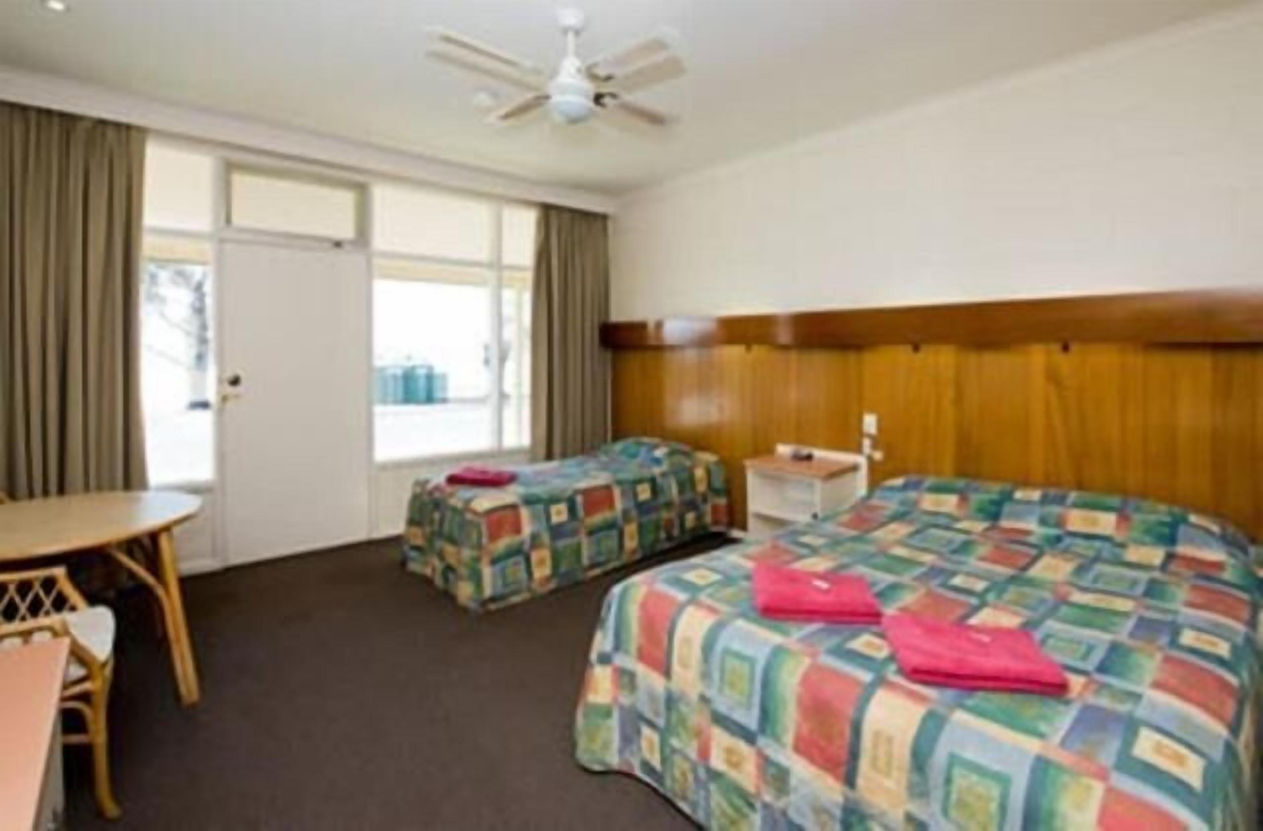 A hotel room with two beds, a wooden headboard, a small table, and a ceiling fan.
