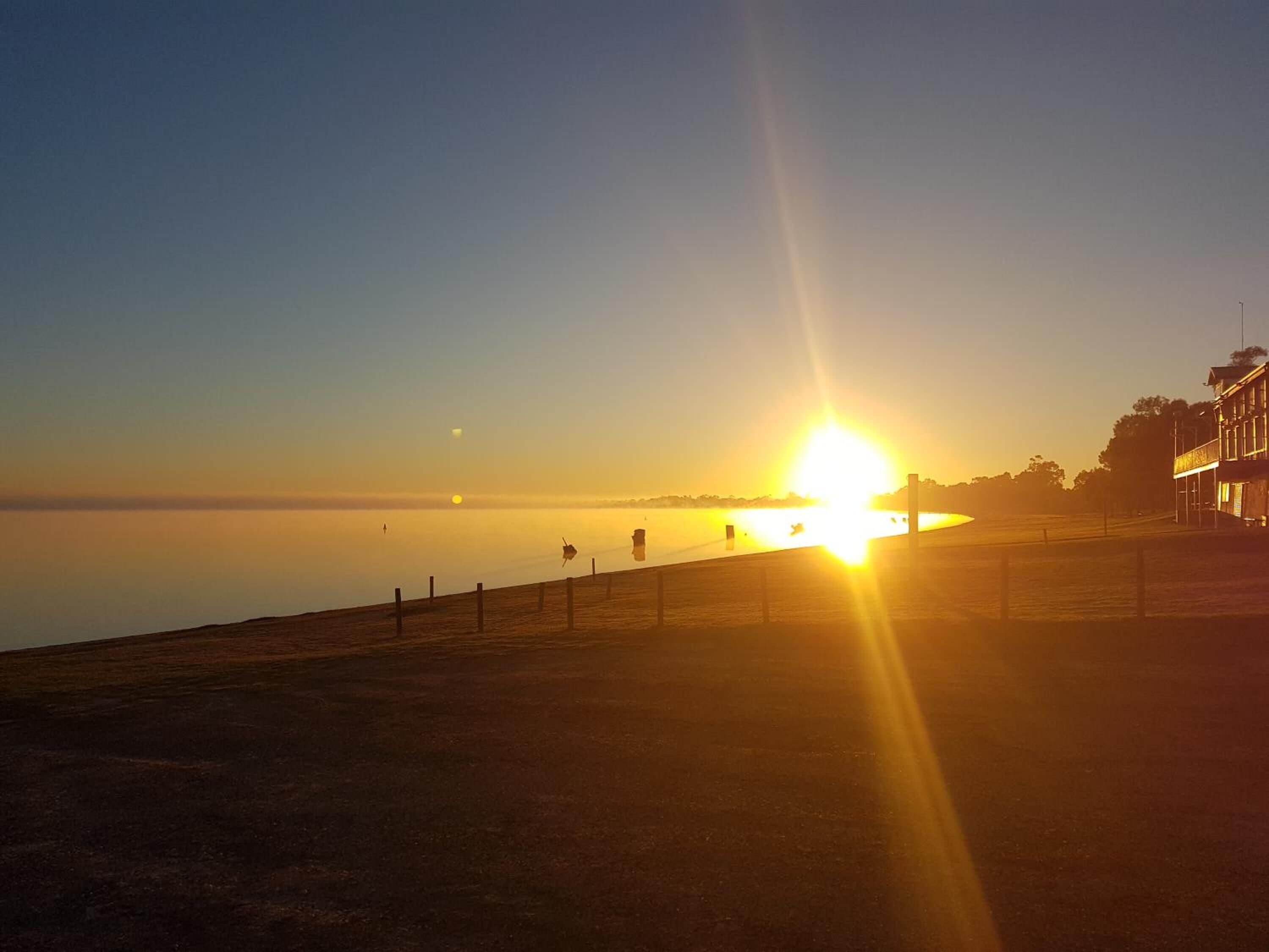 Ein Sonnenuntergang über ruhigem Wasser, im Hintergrund ein Zaun und Gebäude.