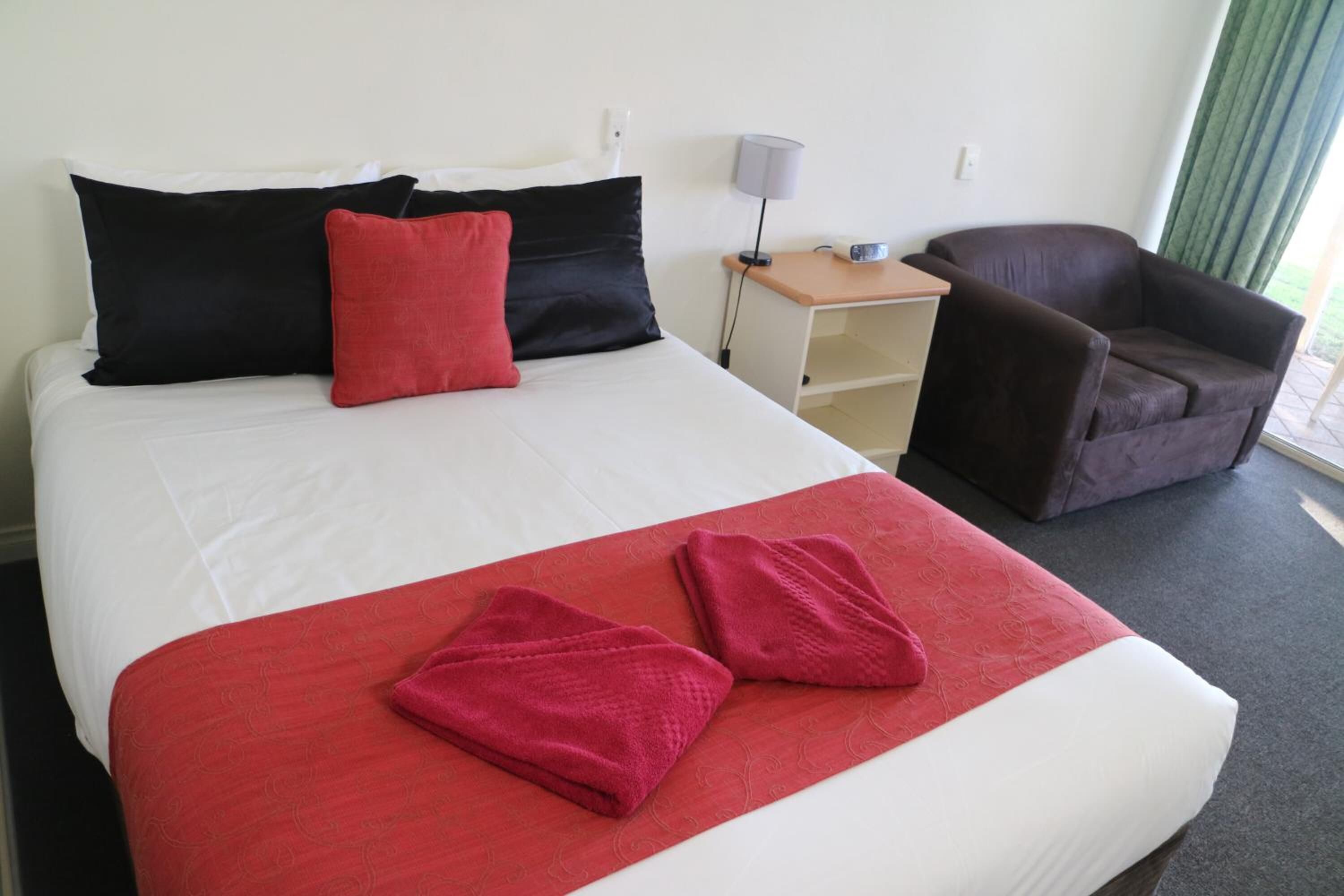 Ein ordentlich bezogenes Bett mit einer roten und weißen Tagesdecke, zwei Kissen und zwei rote Handtücher. Ein brauner Sessel und ein kleiner Tisch mit einer Lampe stehen neben dem Bett.