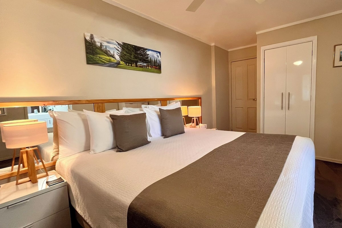 A hotel room with a bed, bedside tables, a TV, a wardrobe, and a painting on the wall.