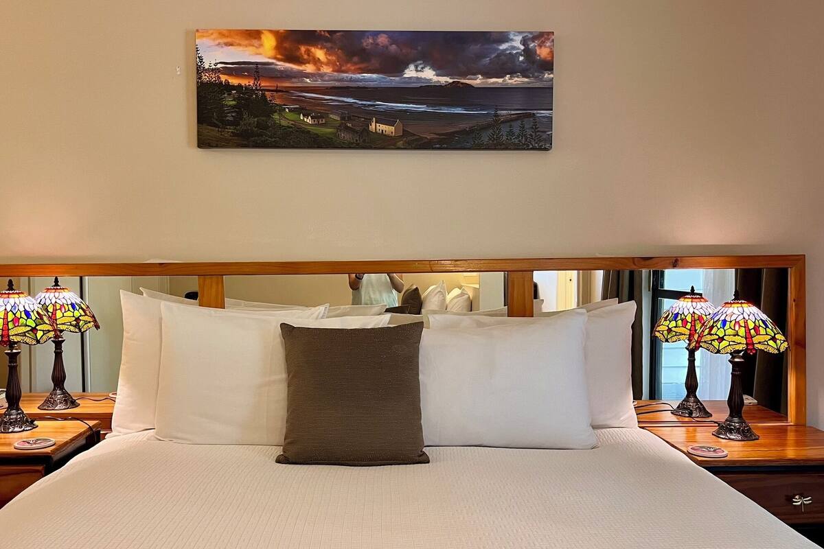 A hotel room with a large bed, two bedside lamps, a painting of a coastal landscape, and a wooden headboard.