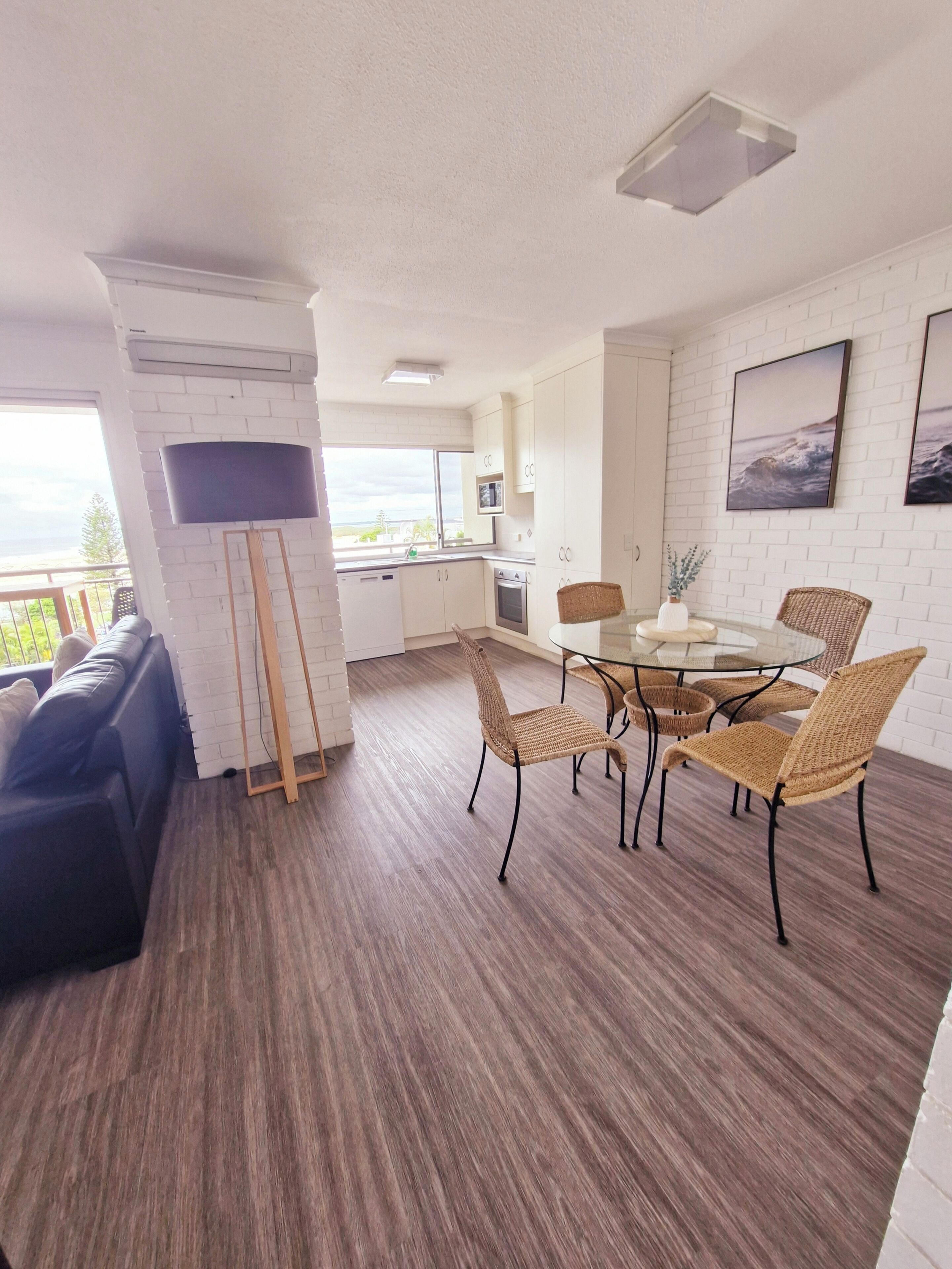 A modern living room with a sofa, a dining table with chairs, a TV mounted on a brick wall, and a kitchen area with cabinets and appliances.