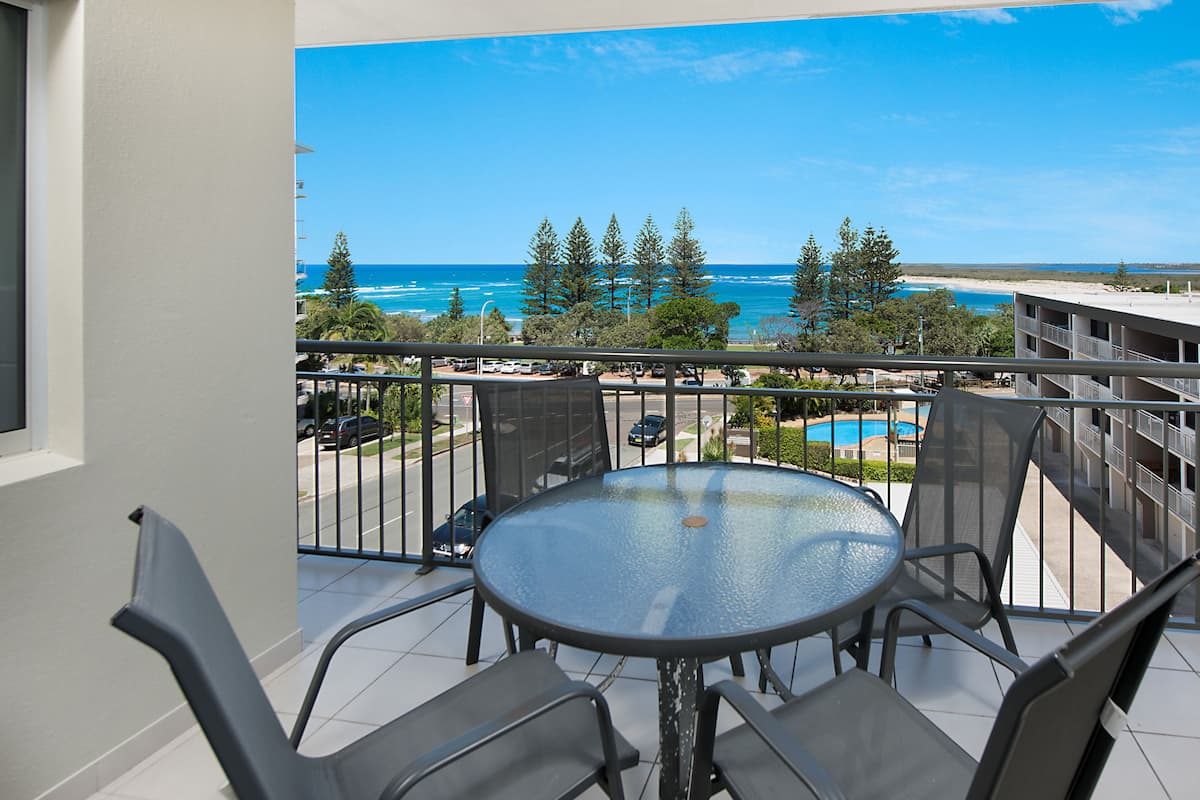 A balcony with a glass table and chairs, overlooking a beach and a pool.
