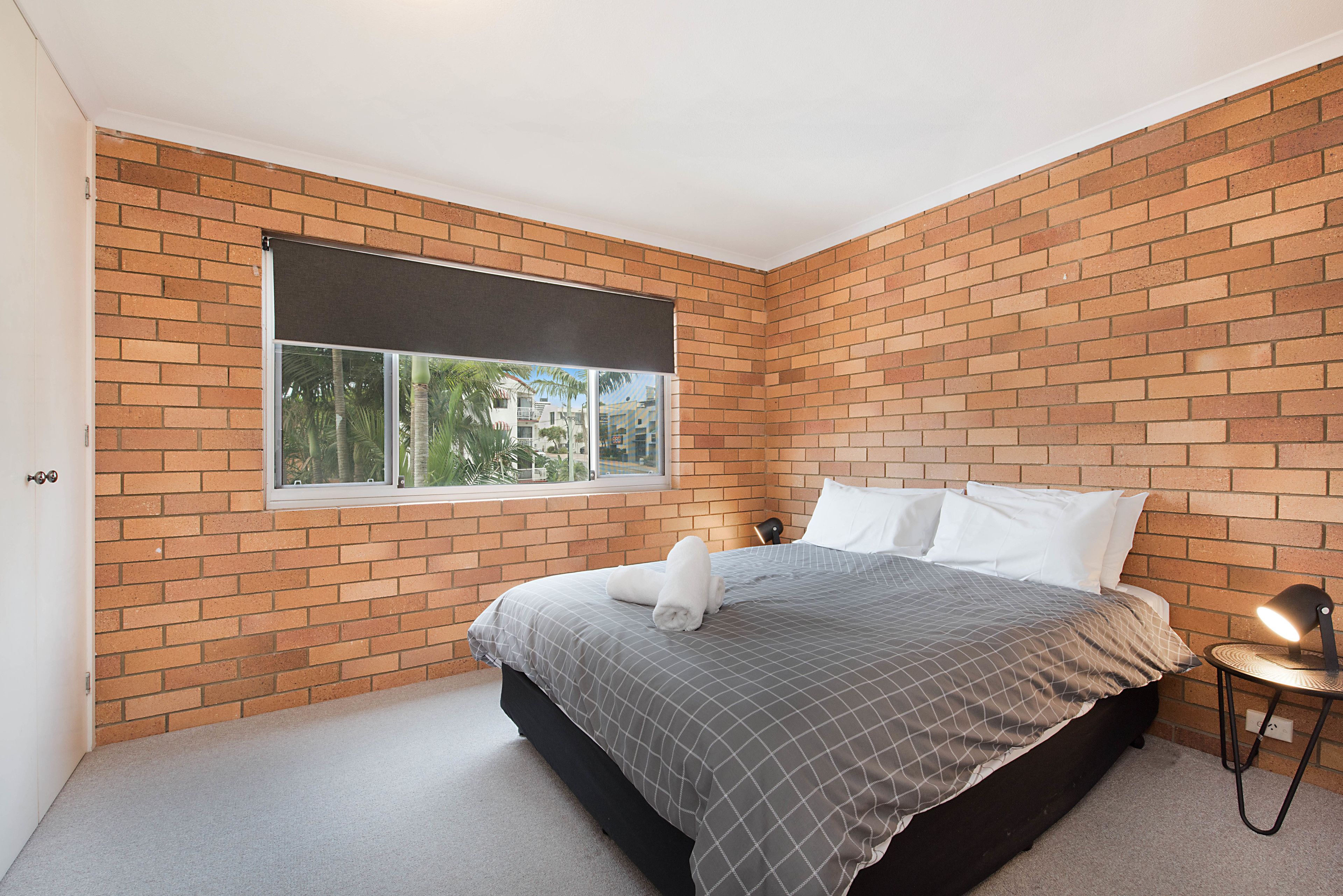 Een slaapkamer met een bakstenen muur, een groot raam, een bed met een grijs geruit sprei, witte kussens en een bedlampje.