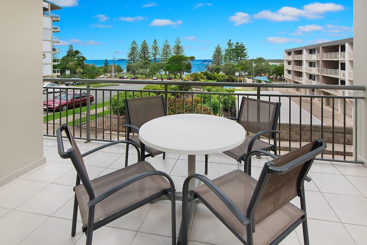 A balcony with a round table and four chairs, overlooking a road with cars and a building with a balcony.