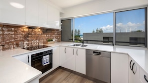 A modern kitchen with white cabinets, a brick accent wall, a dishwasher, and a large window with a view of the sea.