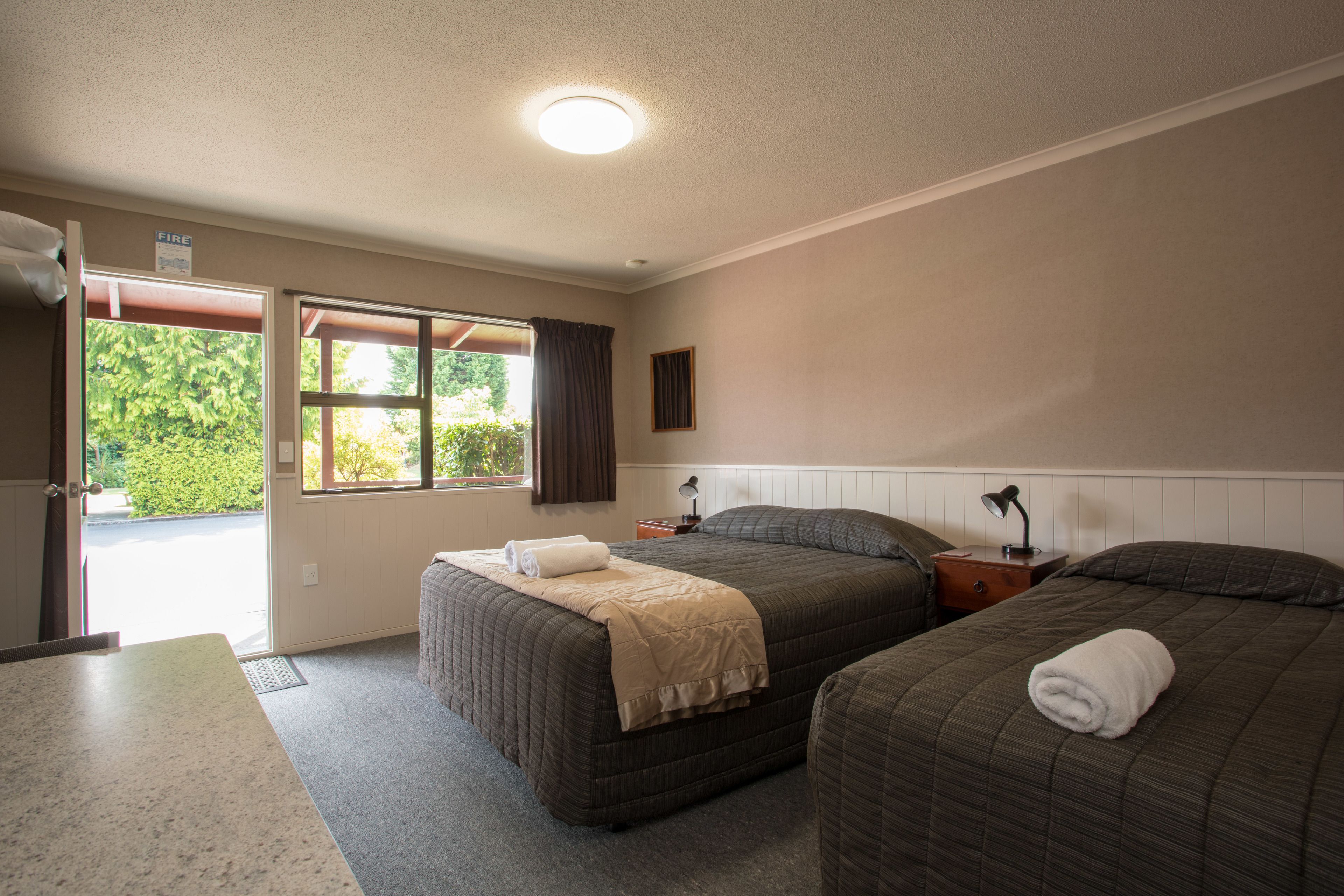 A hotel room with two beds, a window with curtains, a bedside table with a lamp, and a door leading outside.