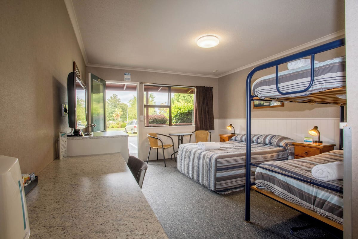 Ein Zimmer mit Stockbetten, einem Schreibtisch, einem Stuhl, einem Tisch, einem Fernseher und einem Fenster mit Blick auf Grünflächen.
