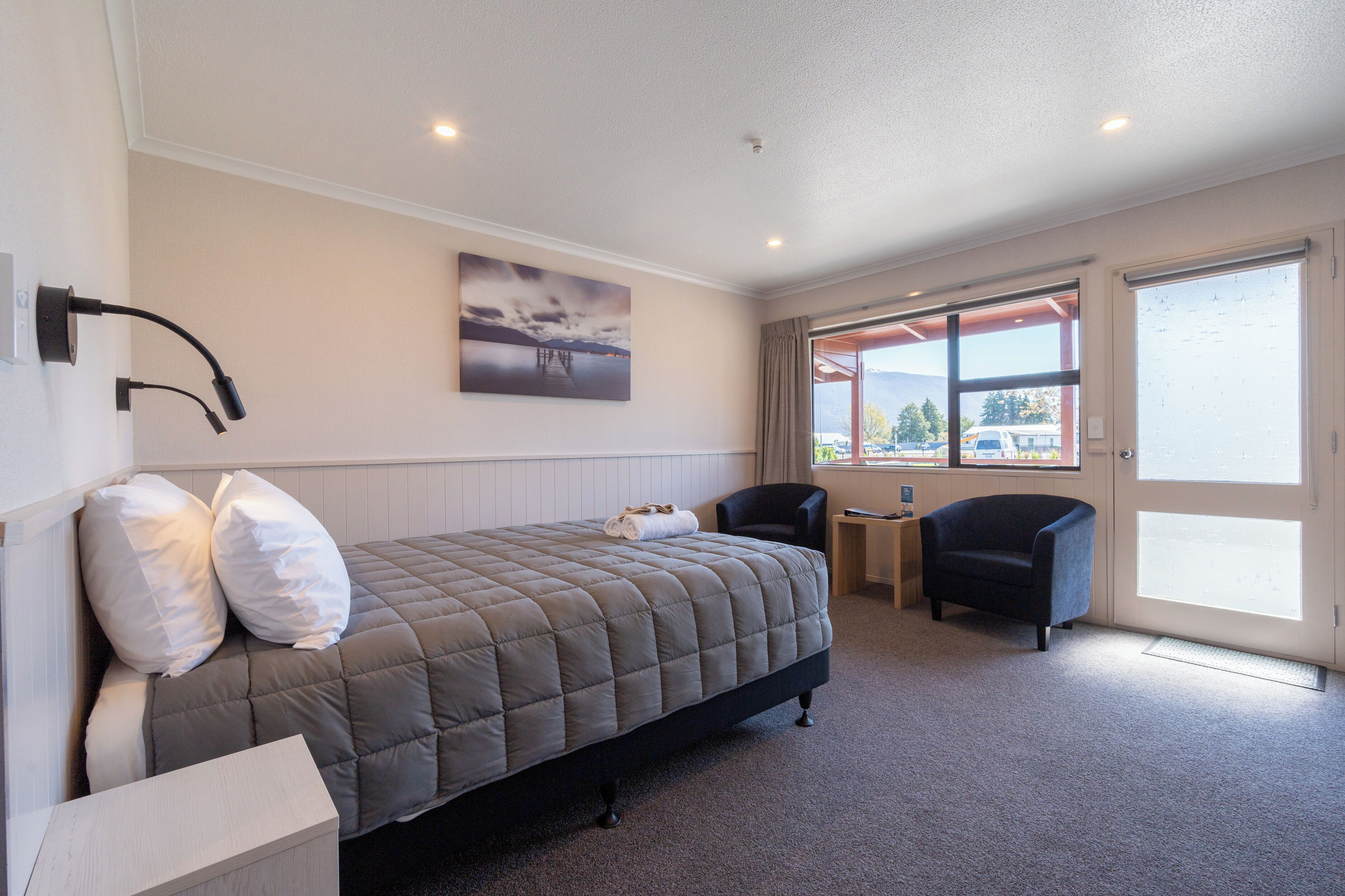 Une chambre à coucher comprenant un lit, une chaise, une petite table et une fenêtre donnant sur un immeuble et des montagnes.
