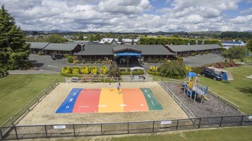 Un parque infantil con una zona de juegos colorida, un edificio identificado como "Te Anau TOP 10 Holiday Park" y un estacionamiento.