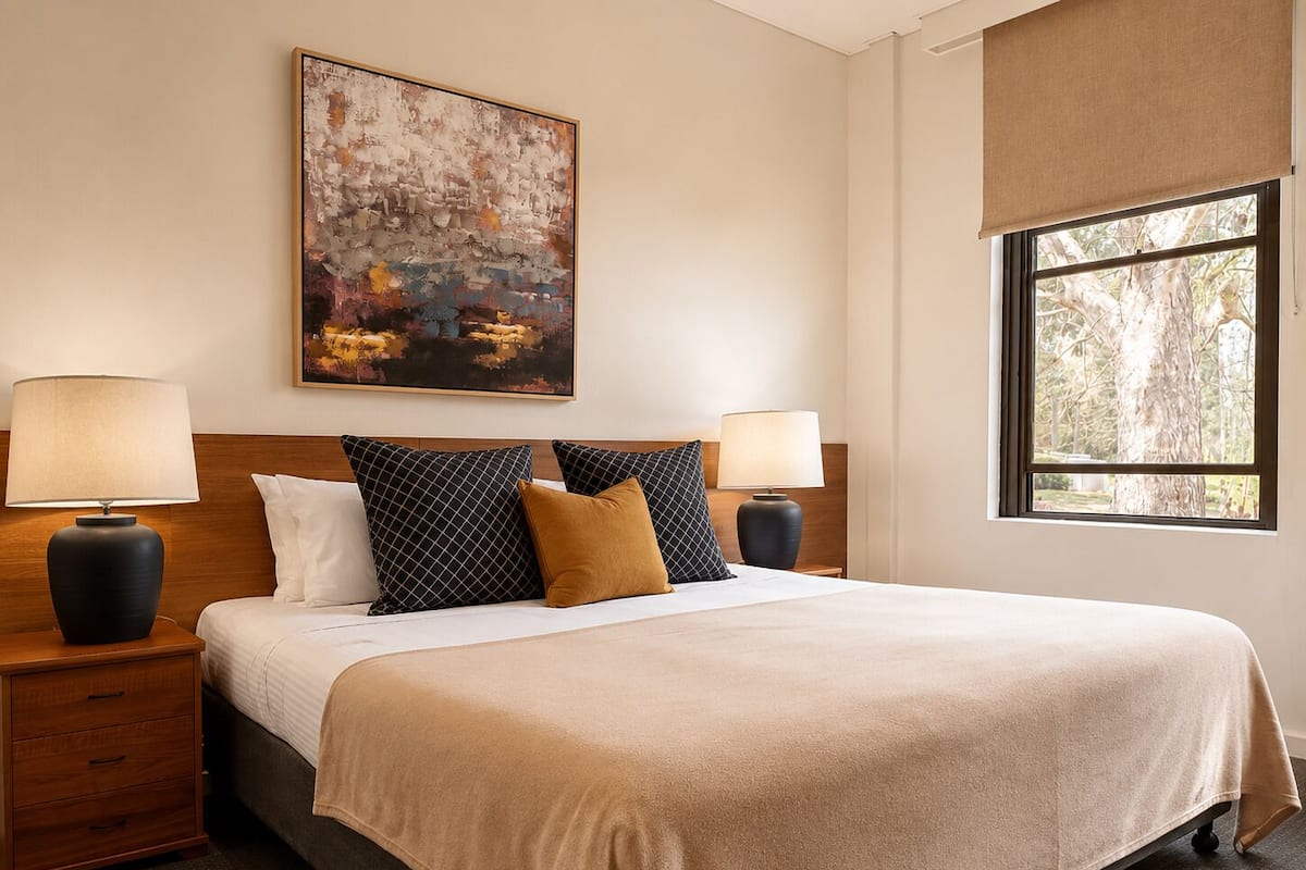 A hotel room with a large bed, two bedside tables with lamps, a large abstract painting on the wall, and a window with a view of trees.
