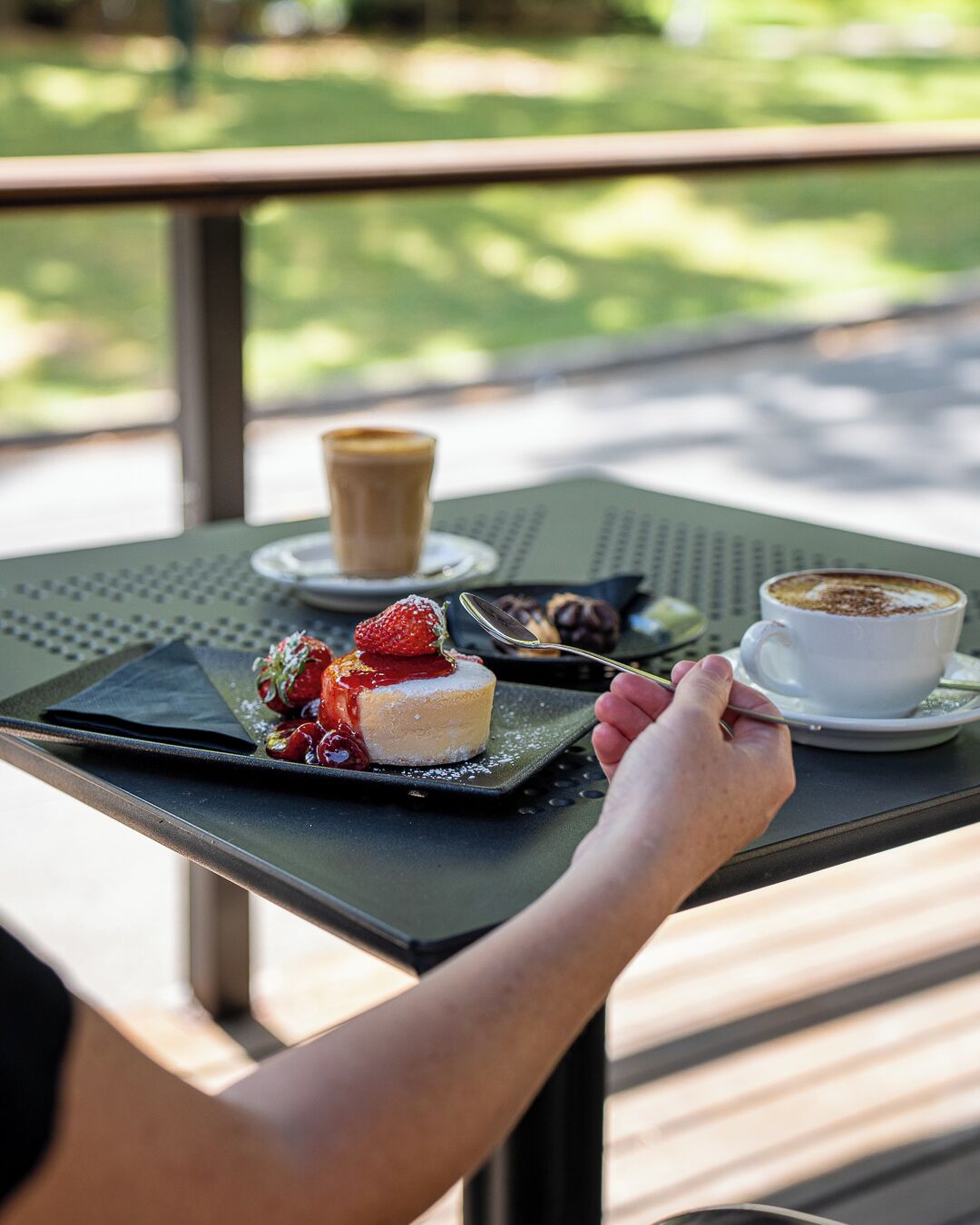 A table with a dessert and two cups of coffee, a person’s hand holding a spoon, and a view of greenery outside.