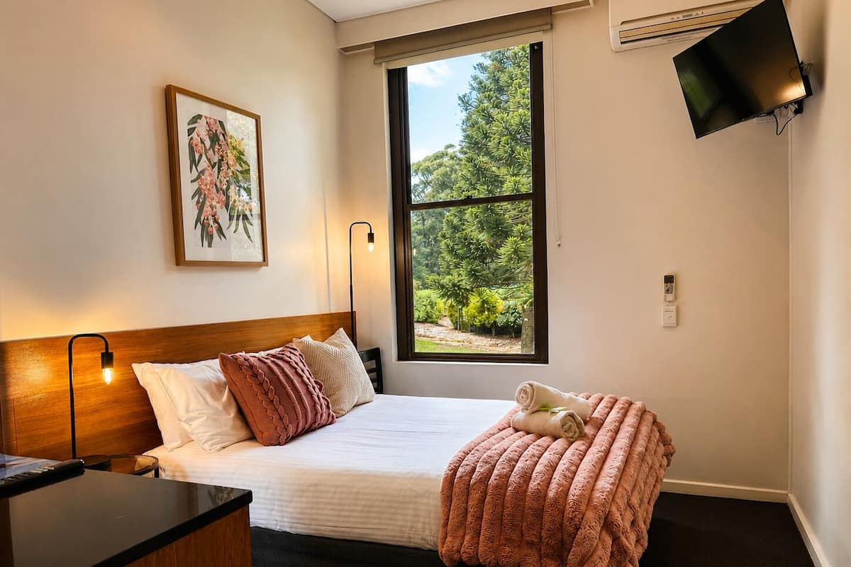 A neatly arranged bedroom with a bed, bedside lamp, and a window with a view of greenery.