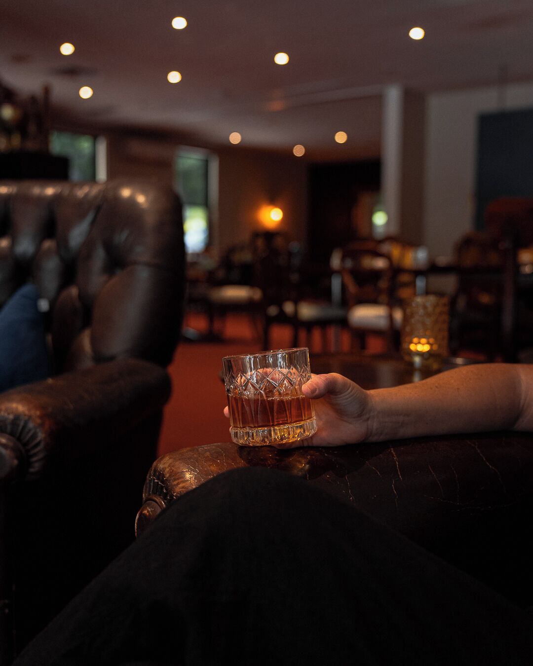 A person holding a glass of whiskey in a dimly lit lounge area with leather seating and ambient lighting.