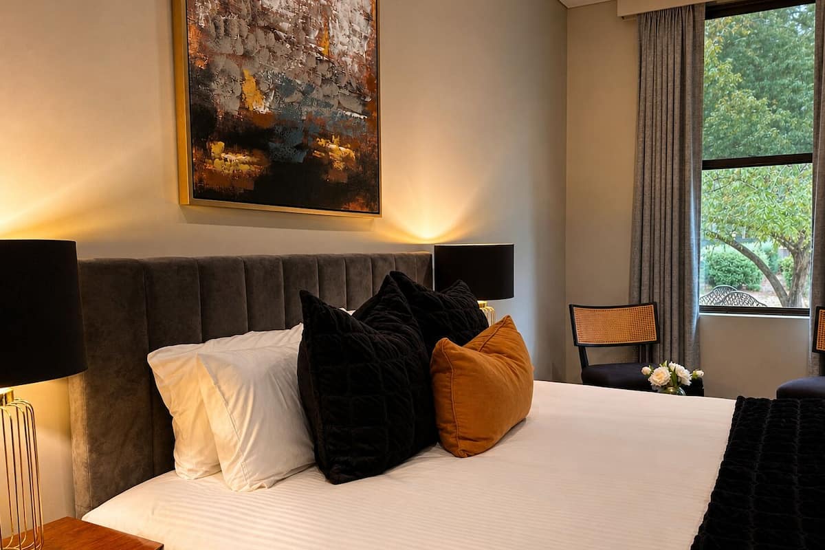 A hotel room with a bed, a nightstand, a lamp, a chair, a window with curtains, and a painting on the wall.