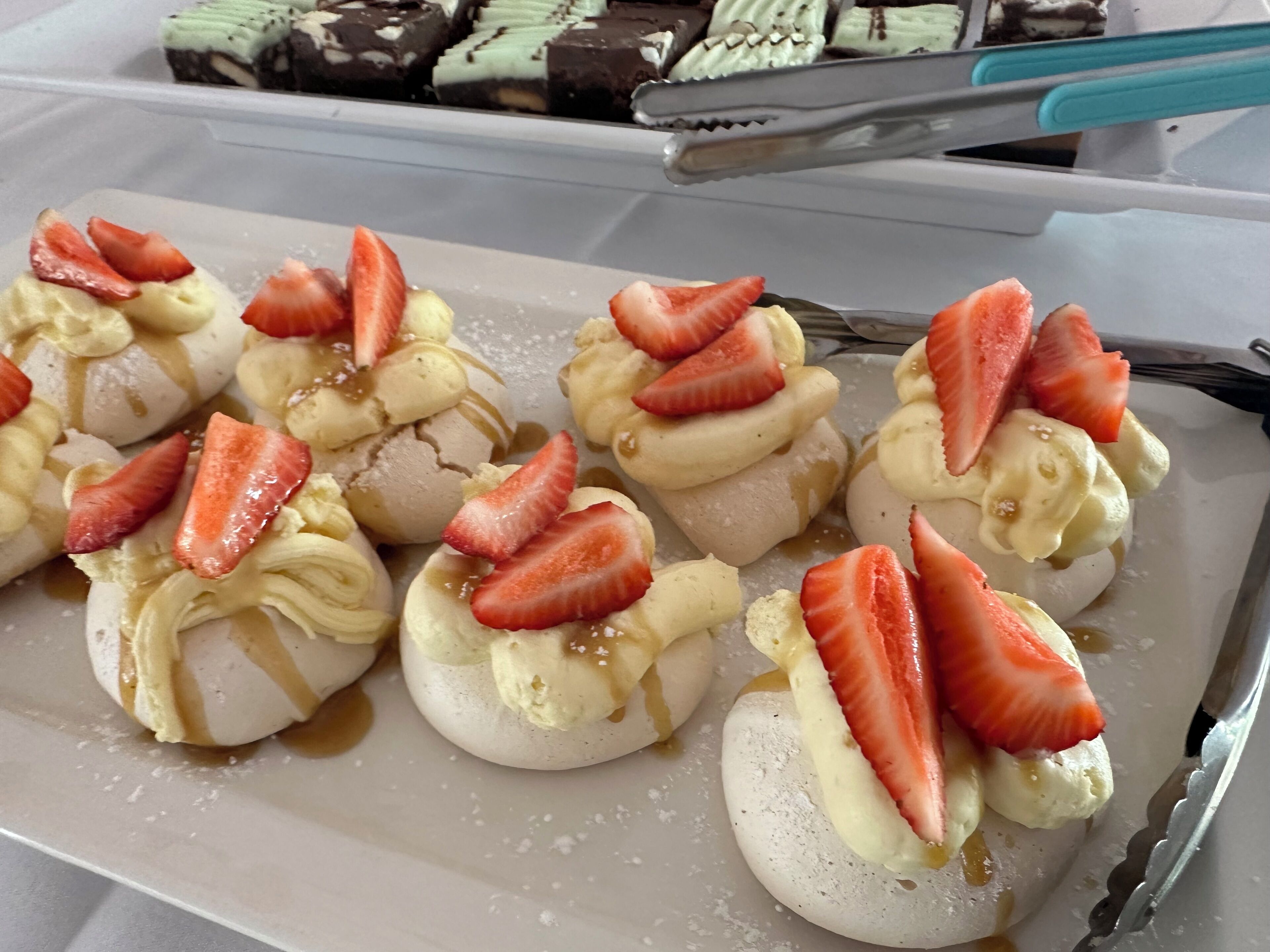 A tray of dessert items topped with strawberries and bananas, with a pair of tongs resting on the tray.