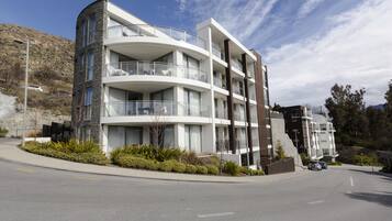 A modern multi-story building with balconies, a landscaped area with shrubs, and a clear road in front.