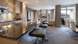 A modern kitchen with a central island, bar stools, a dining area with chairs, and a large window offering a mountain view. - Queenstown Village Apartments (Kawarau Falls)