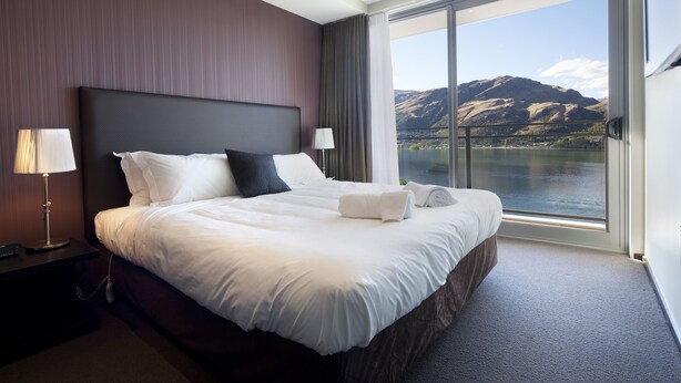 A hotel room with a large bed, two bedside lamps, a view of mountains and a lake through the window. - Queenstown Village Apartments (Kawarau Falls)