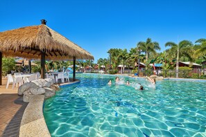 Uma piscina azul límpida com uma cobertura de palha, rodeada por palmeiras e espreguiçadeiras.