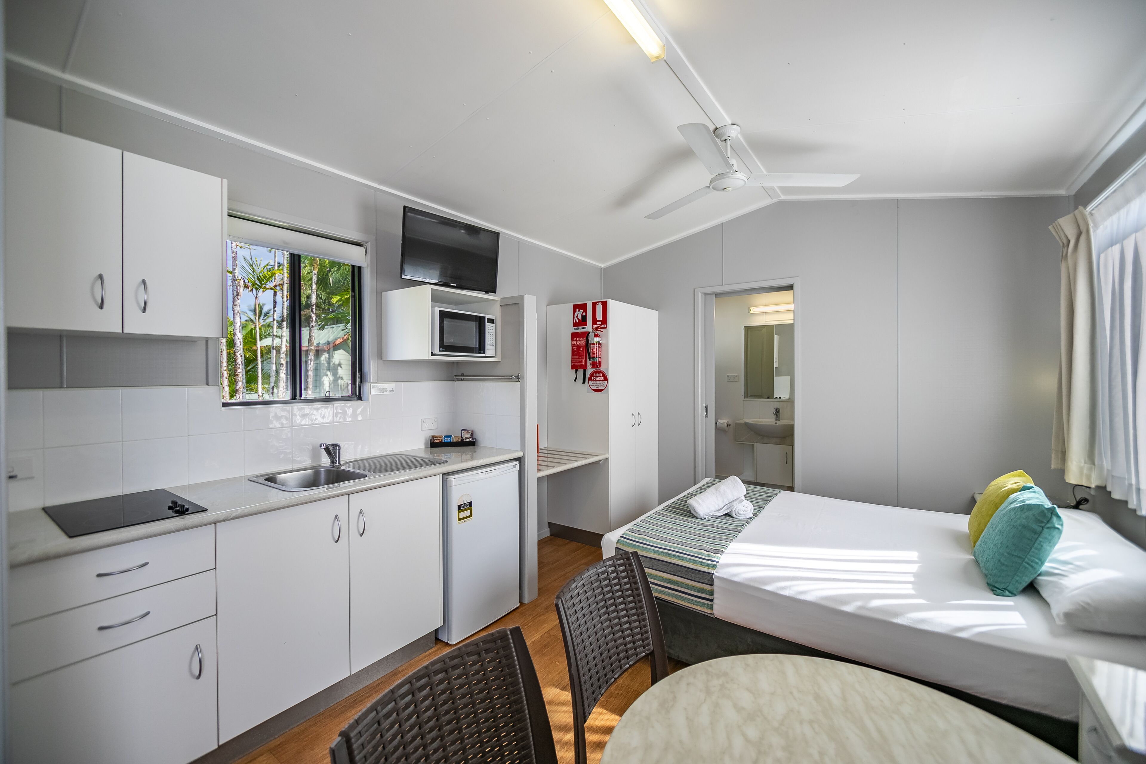 A compact hotel room with a kitchenette, a bed with a striped bedspread, a ceiling fan, and a view of the outdoors.