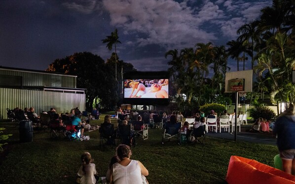 Exibição de cinema ao ar livre com uma tela grande, pessoas sentadas em cadeiras de gramado e uma quadra de basquete ao fundo.