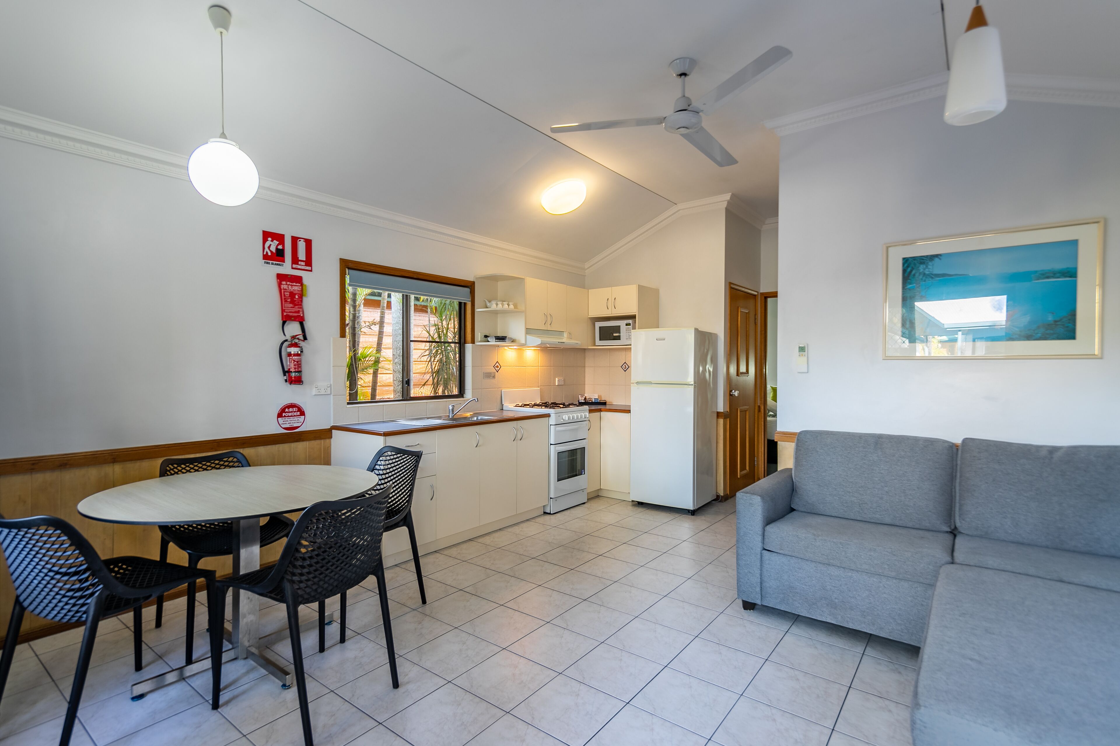 Une cuisine moderne avec des meubles blancs, une table à manger avec des chaises, un canapé gris et un ventilateur de plafond.