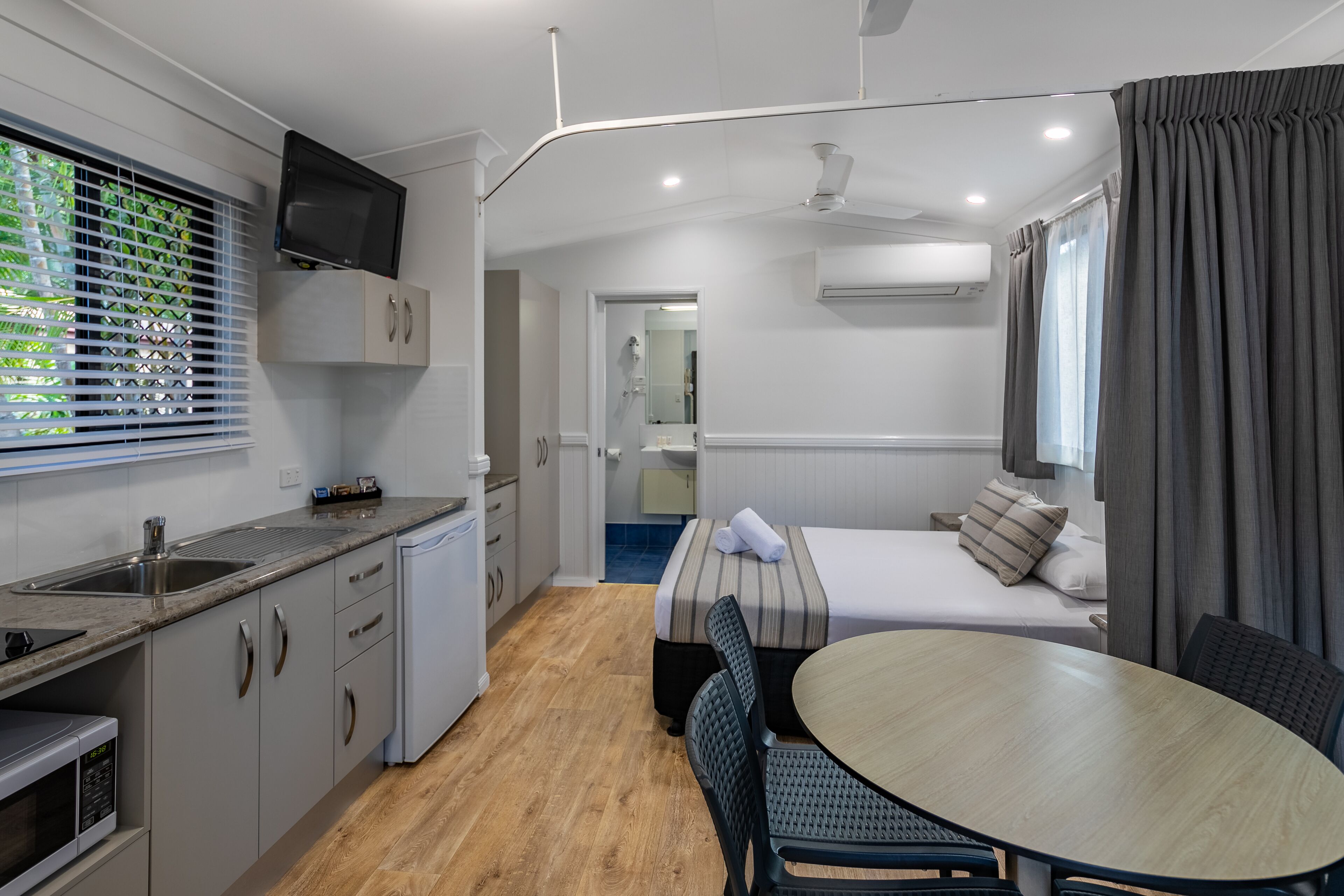 A compact hotel room with a kitchenette, a bed with a striped bedspread, a dining table with chairs, and a TV mounted above the sink.