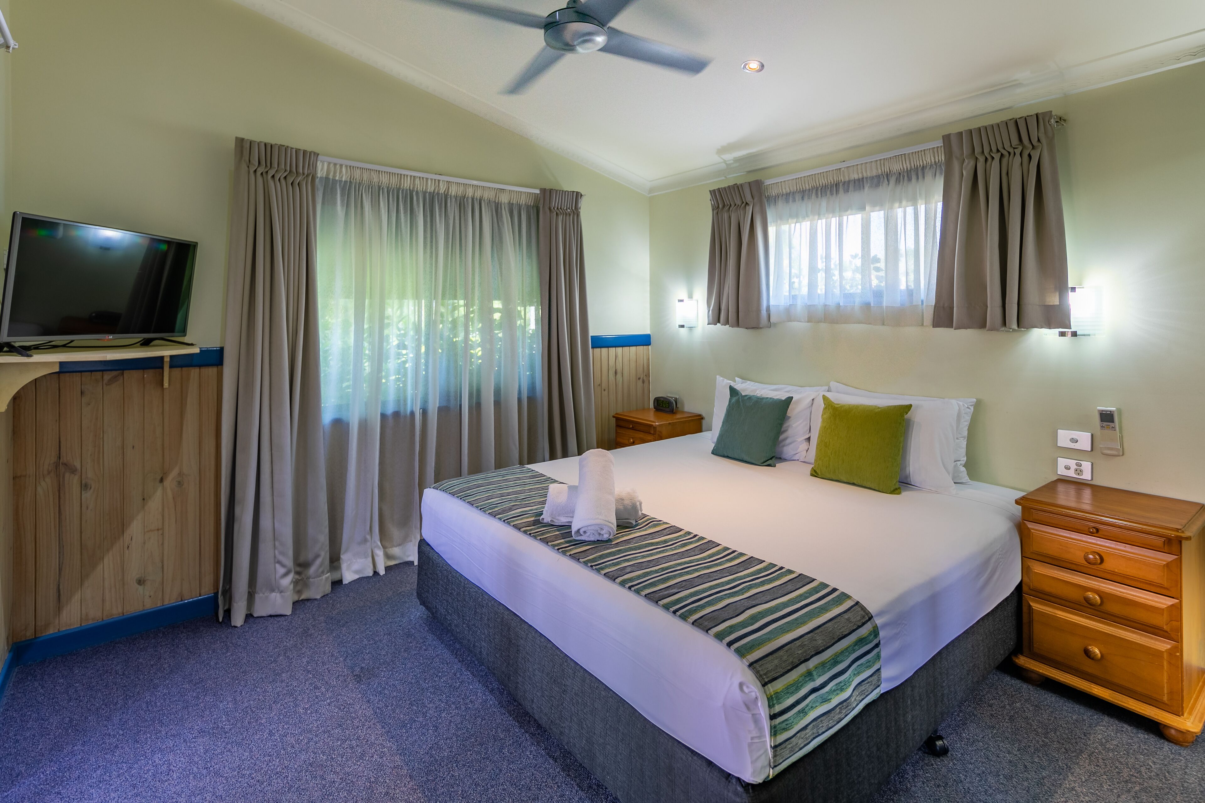 Habitación de hotel con una cama, un televisor, una mesita de noche de madera y un ventilador de techo.