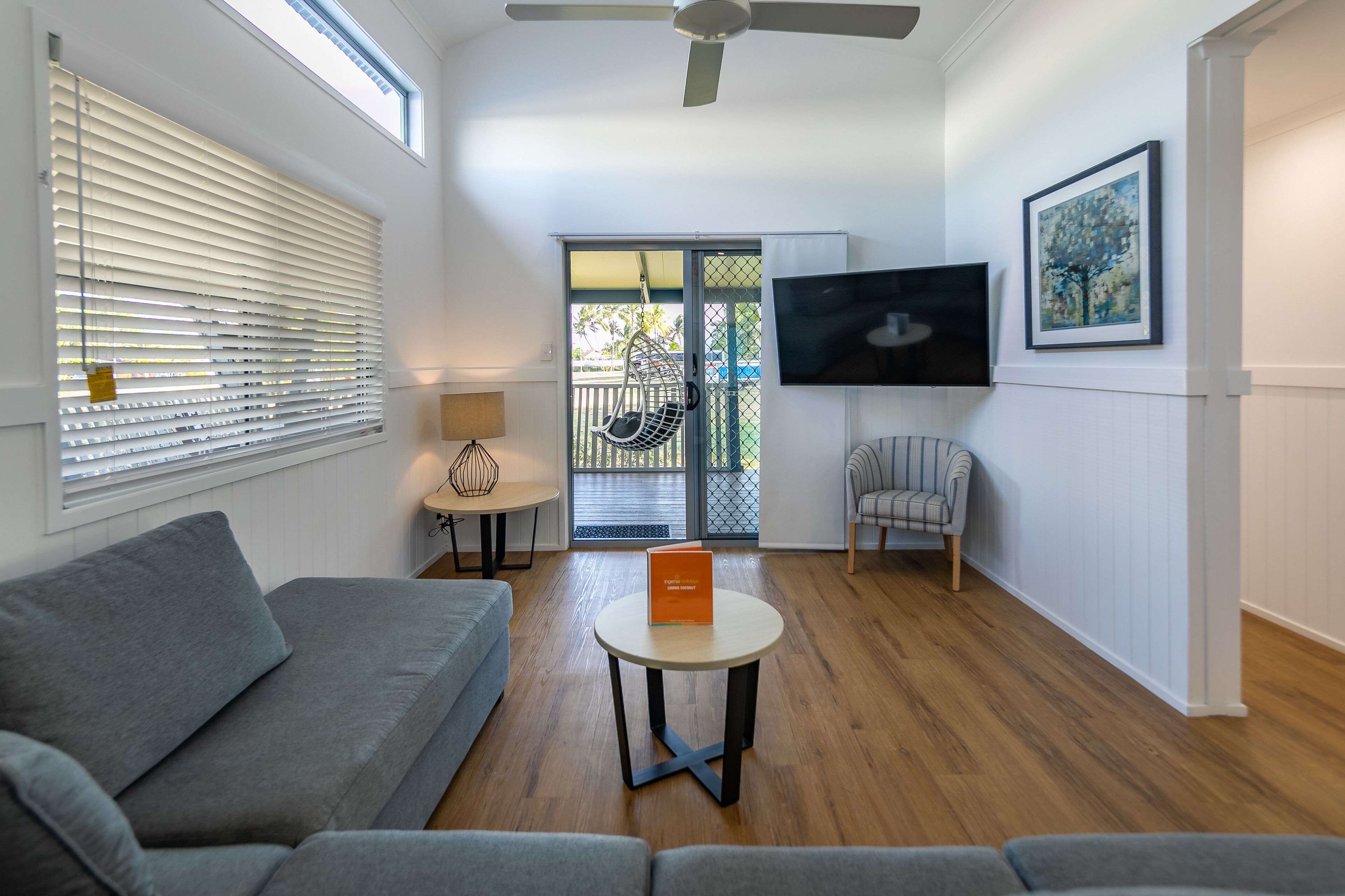 A living room with a grey sofa, a round coffee table with an orange book, a TV on the wall, and a hanging chair near a sliding glass door.