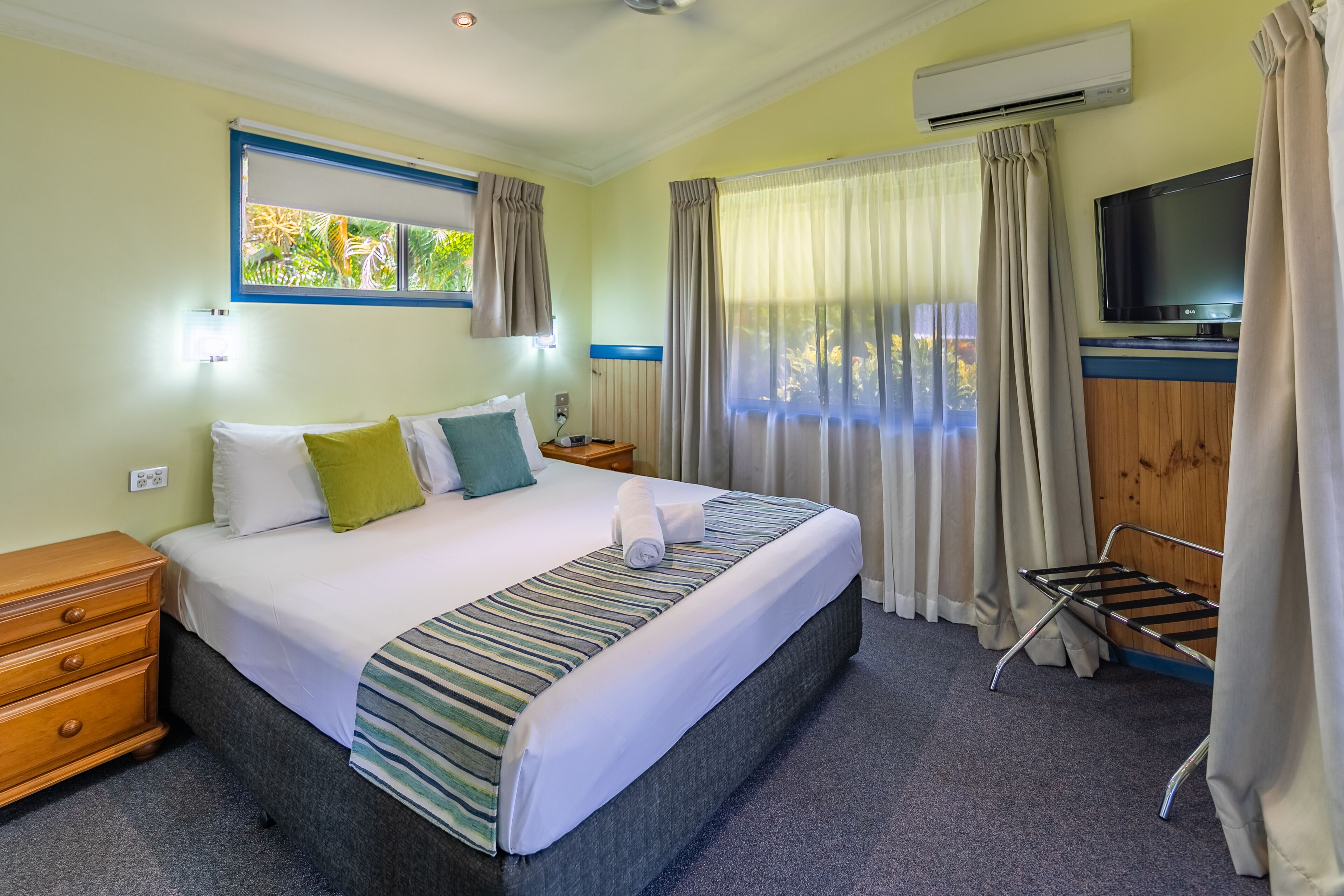 A hotel room with a large bed, a TV, a window with curtains, a wooden nightstand, and a small table.
