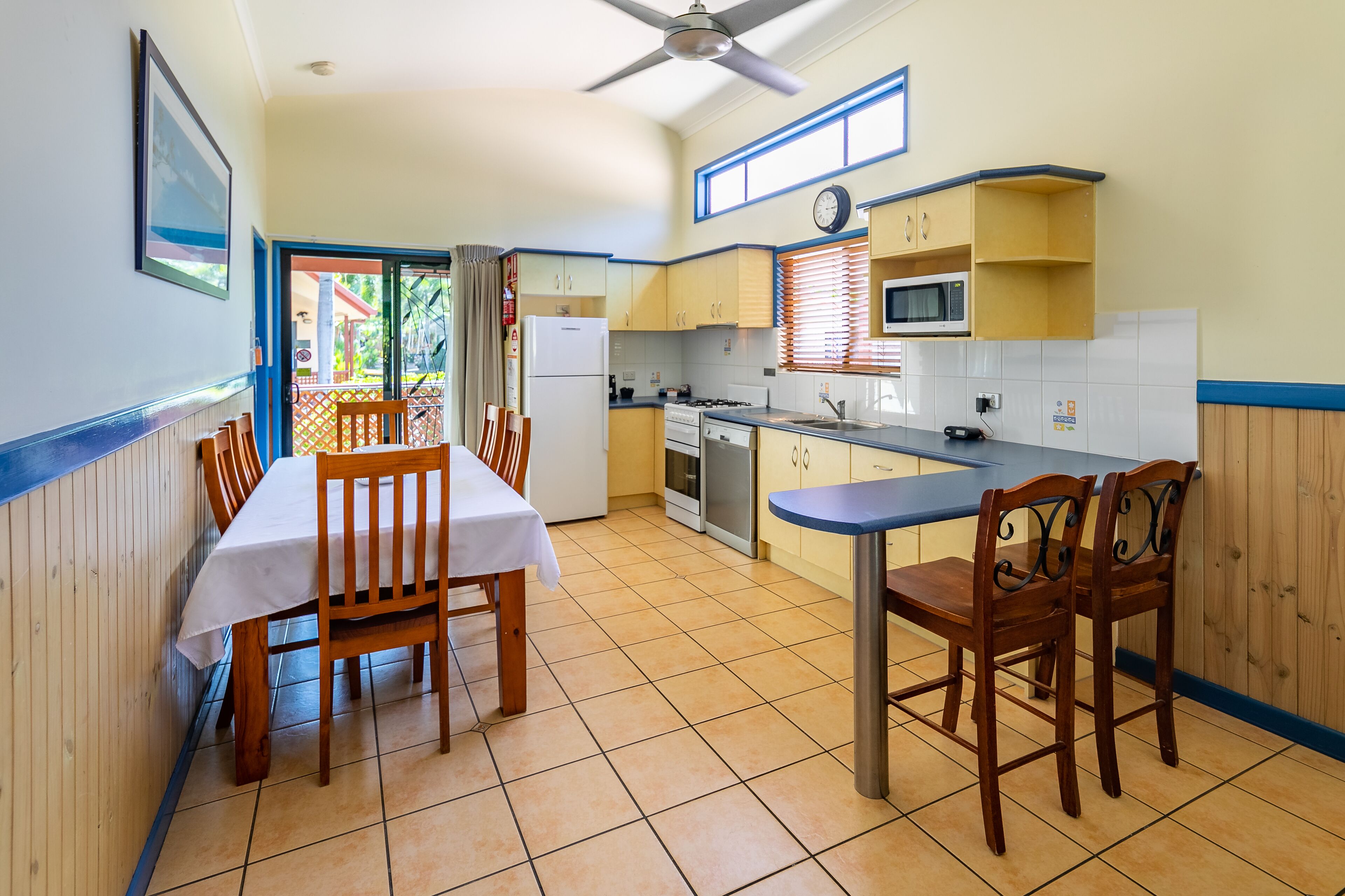 A compact kitchen with a dining area, featuring a refrigerator, stove, microwave, and wooden chairs.