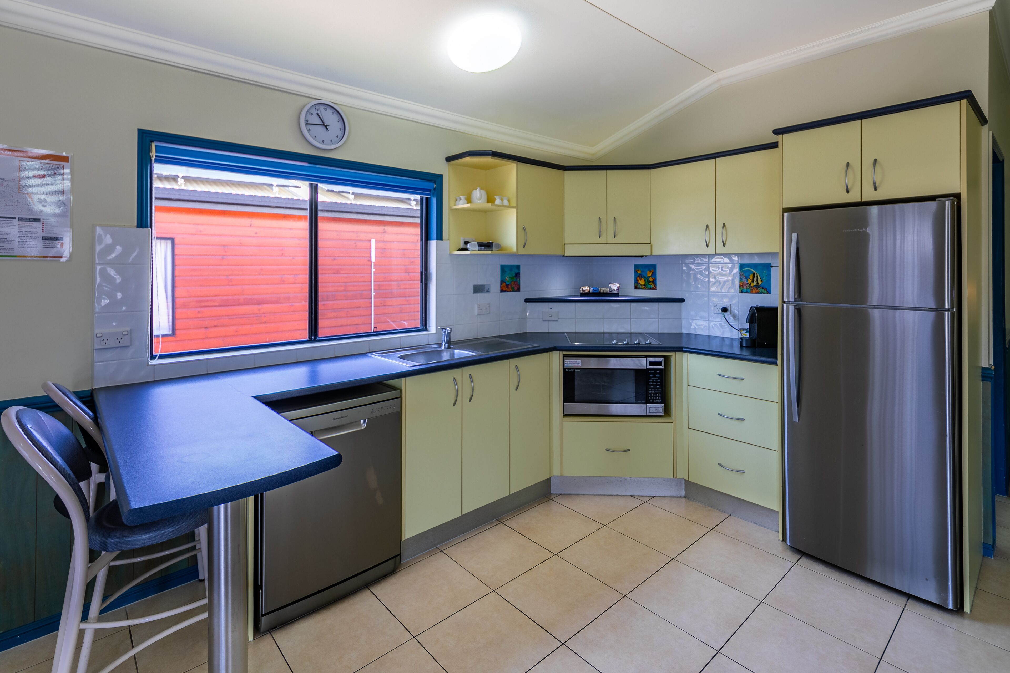 A kitchen with a stainless steel refrigerator, a microwave, and a dishwasher. There is a window with red shutters and a clock on the wall.