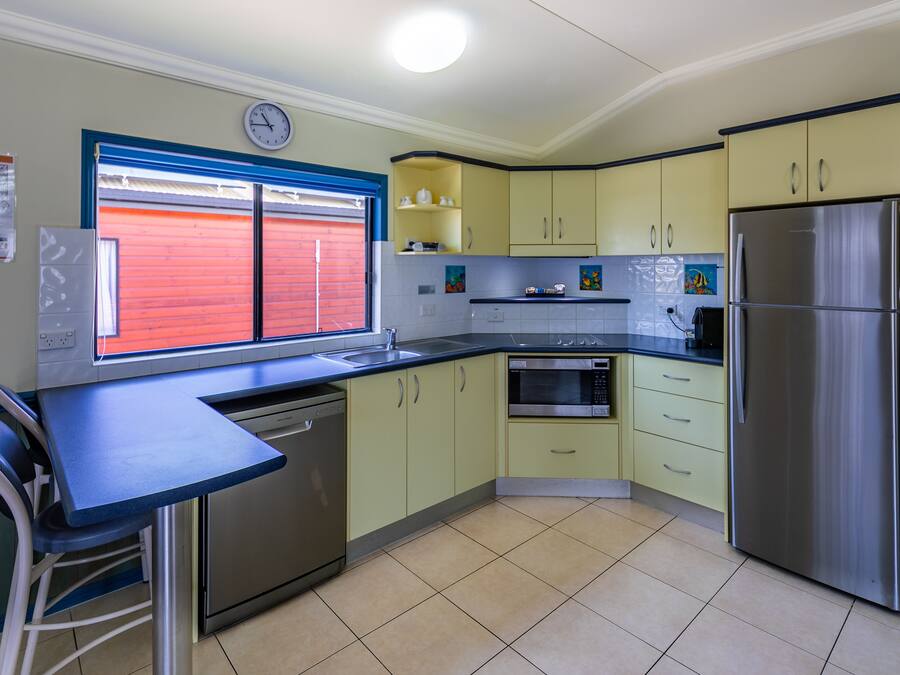 A kitchen with a stainless steel refrigerator, a microwave, and a dishwasher. There is a window with red shutters and a clock on the wall.