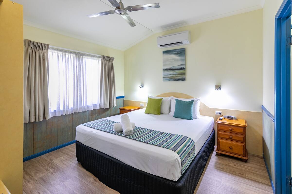 Habitación de hotel con cama, ventilador de techo, aire acondicionado, mesita de noche y un cuadro en la pared.