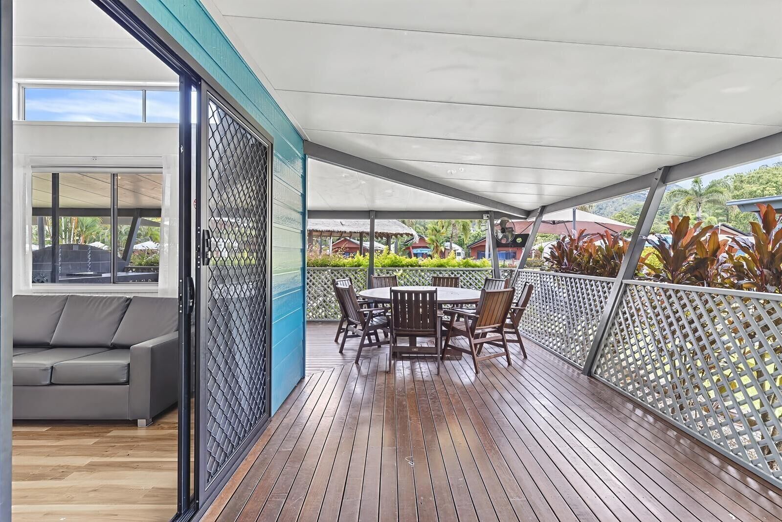 Un patio exterior con terraza de madera, una mesa redonda con sillas y vistas a un jardín exuberante y edificios al fondo.