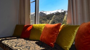 A bed with a patterned bedspread and colorful pillows, a window with a view of a snow-covered mountain and forest. - Squatter's Run Apartments (Thredbo)