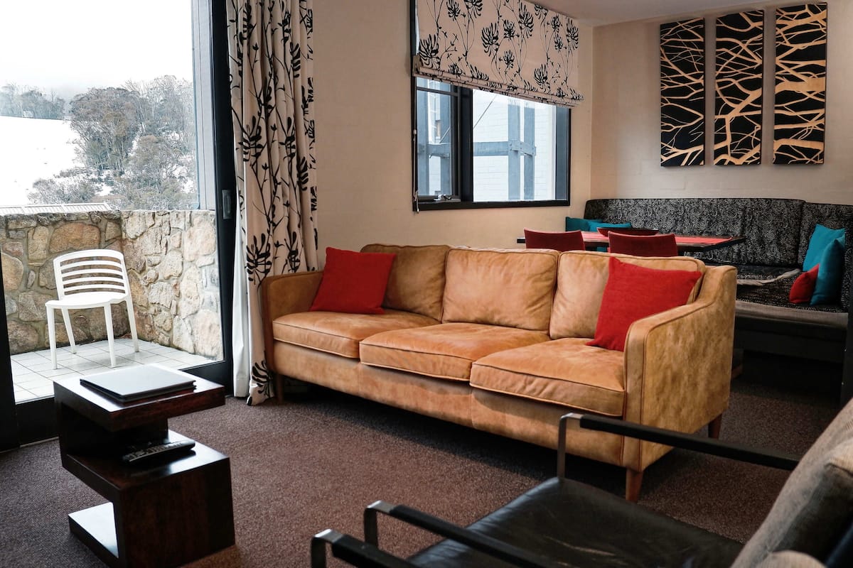 A living room with a leather sofa, a white chair, and a coffee table. There is a window with a view of trees and a stone wall.